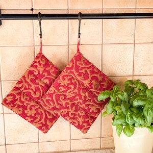 May include: Two red and gold patterned pot holders hanging on hooks. The pot holders have a quilted design and are made of fabric.