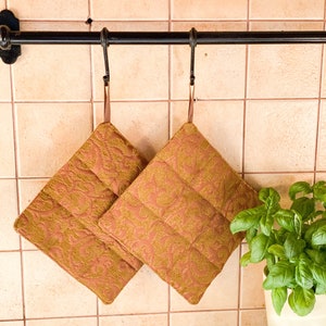 Op de afbeelding: Twee bruine en gouden gewatteerde ovenwanten met een bloemenpatroon hangen aan een zwarte metalen rek tegen een beige betegelde muur.