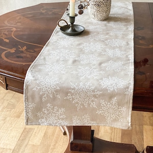 Op de afbeelding: Een beige tafelloper met een wit sneeuwvlokpatroon ligt op een donkere houten tafel. Een brandende kaars in een messing houder en een decoratieve vaas met takken en dennenappels staan op de loper. De tafel heeft een ingelegd ontwerp.