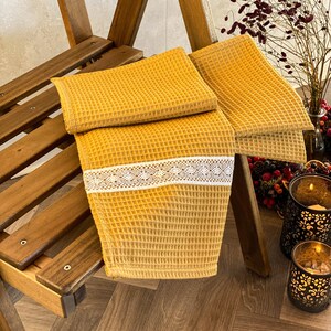 Puede incluir: Dos toallas de cocina de tejido de waffle de color amarillo mostaza con ribete de encaje blanco están dobladas y apiladas en una silla de madera. Las toallas están sobre un suelo de madera con un patrón de espiga.