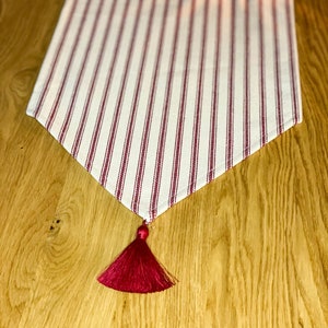 Puede incluir: Un camino de mesa a rayas blancas y rojas con una borla roja. El camino de mesa está sobre una mesa de madera.