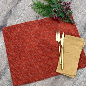 Könnte beinhalten: Ein roter Tischset mit Rautenmuster, goldene Gabel und Messer sowie eine gefaltete beige Serviette. Ein Zweig mit roten Beeren liegt auf der Holzoberfläche, was eine festliche Tischdekoration ergibt.