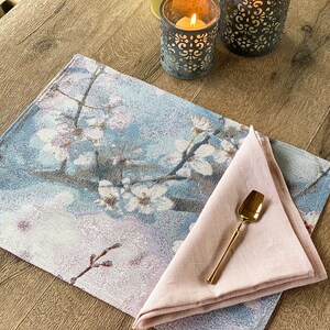 May include: A blue and white floral placemat with a pink linen napkin and a gold spoon on top. The placemat has a design of white flowers on a blue background. The napkin is folded in a triangle shape. The spoon is lying on top of the napkin.