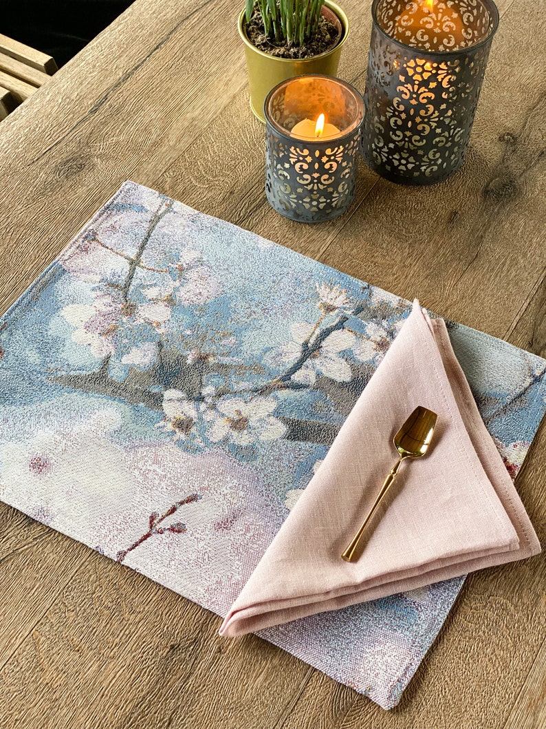 May include: A blue and white floral placemat with a pink linen napkin and a gold spoon on top. The placemat has a design of white flowers on a blue background. The napkin is folded in a triangle shape. The spoon is lying on top of the napkin.