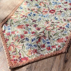 Pode incluir: Um corredor de mesa bege com um padr&atilde;o floral em tons de vermelho, azul e verde. O corredor tem uma borda de croch&ecirc; marrom.