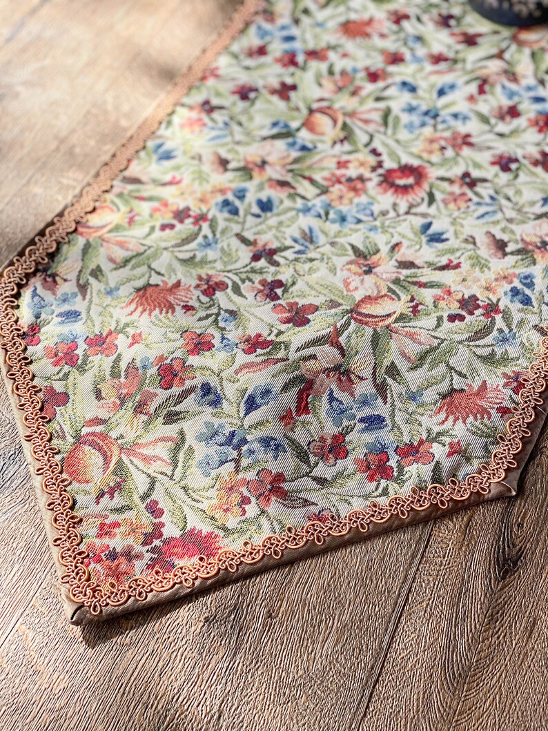 Pode incluir: Um corredor de mesa bege com um padr&atilde;o floral em tons de vermelho, azul e verde. O corredor tem uma borda de croch&ecirc; marrom.