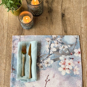 May include: A blue and white floral placemat with a light blue linen napkin, a gold fork and knife, and two decorative candle holders with lit candles on a wooden surface.