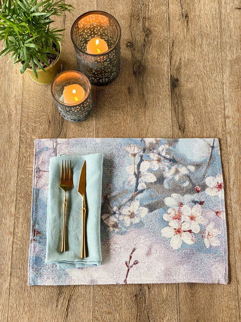 May include: A blue and white floral placemat with a light blue linen napkin, a gold fork and knife, and two decorative candle holders with lit candles on a wooden surface.