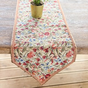 Pode incluir: Um corredor de mesa floral com fundo bege e uma delicada renda cor de p&ecirc;ssego. O corredor apresenta um padr&atilde;o repetitivo de flores e folhas vermelhas, rosas e azuis.
