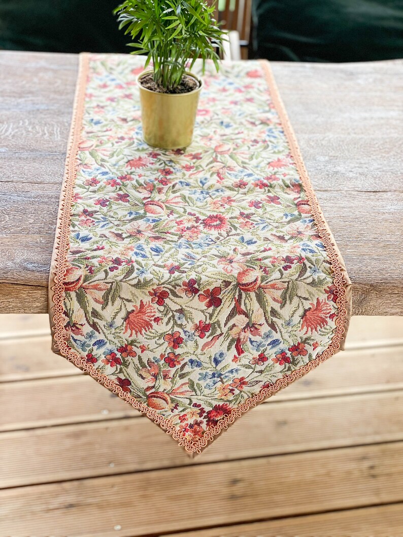 Pode incluir: Um corredor de mesa floral com fundo bege e uma delicada renda cor de p&ecirc;ssego. O corredor apresenta um padr&atilde;o repetitivo de flores e folhas vermelhas, rosas e azuis.