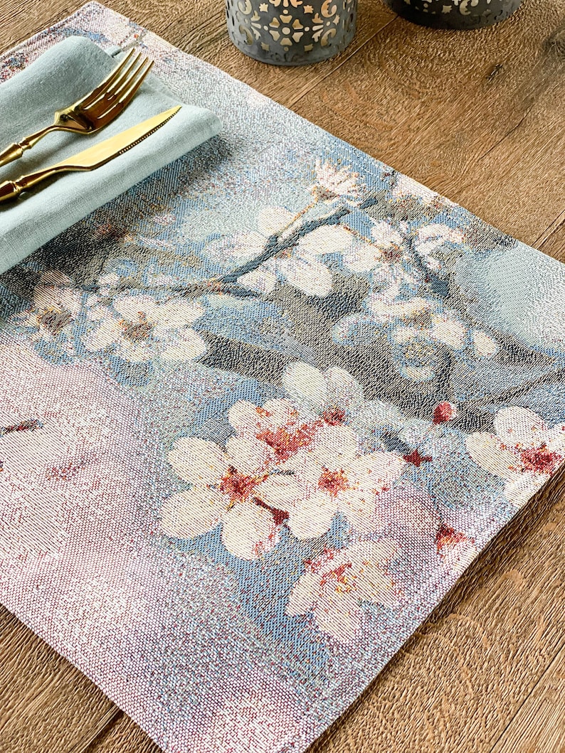 May include: A blue and white placemat with a floral design featuring white flowers with pink centers. The placemat is made of woven fabric and has a textured surface.