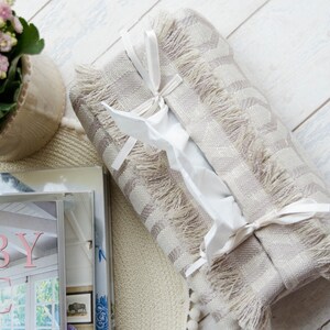 Peut inclure: Un couvre-bo&icirc;te &agrave; mouchoirs ray&eacute; beige et blanc avec une bordure en franges et des liens en ruban blanc. Le couvre-bo&icirc;te est pos&eacute; sur une pile de livres avec le titre "The Abby Chic Home" visible.