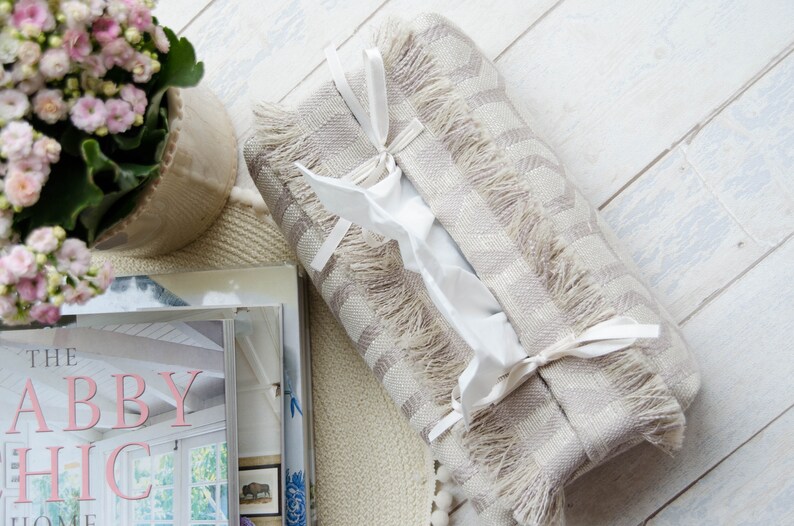Peut inclure: Un couvre-bo&icirc;te &agrave; mouchoirs ray&eacute; beige et blanc avec une bordure en franges et des liens en ruban blanc. Le couvre-bo&icirc;te est pos&eacute; sur une pile de livres avec le titre "The Abby Chic Home" visible.