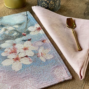 May include: A blue and pink woven placemat with a floral design. A pink linen napkin is folded on top of the placemat, with a gold-colored serving spoon on top of the napkin.