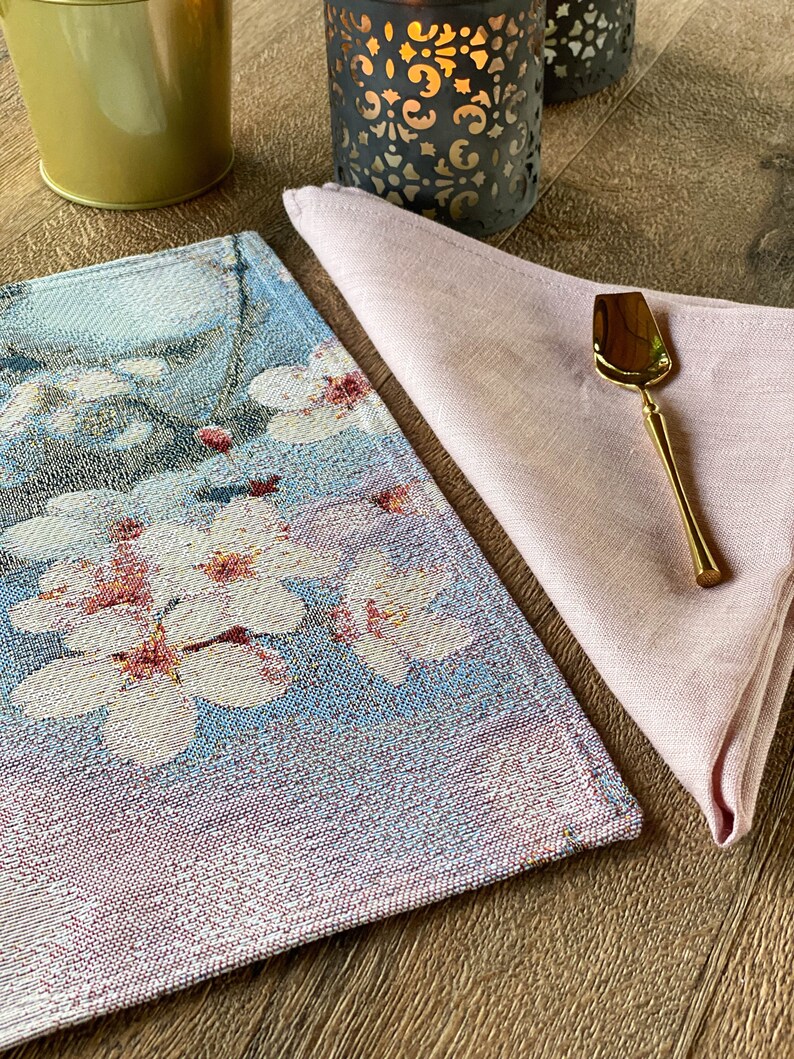 May include: A blue and pink woven placemat with a floral design. A pink linen napkin is folded on top of the placemat, with a gold-colored serving spoon on top of the napkin.