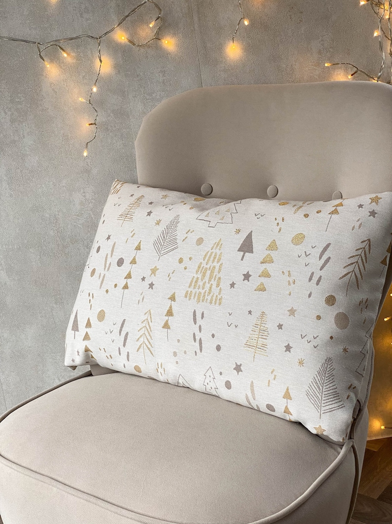 May include: A white throw pillow with a gold and brown pattern of stylized trees, stars, and dots. The pillow is on a beige armchair.