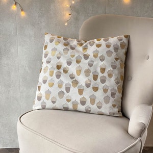May include: A cream-colored armchair with a throw pillow featuring a pattern of gold and brown acorns. The chair is in front of a wall with a string of fairy lights.