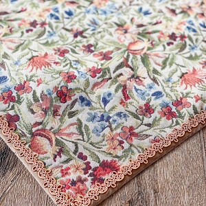 Pode incluir: Um corredor de mesa floral com uma borda de renda bege. O corredor de mesa apresenta um fundo branco com um padr&atilde;o repetitivo de flores vermelhas, rosas, azuis e verdes.