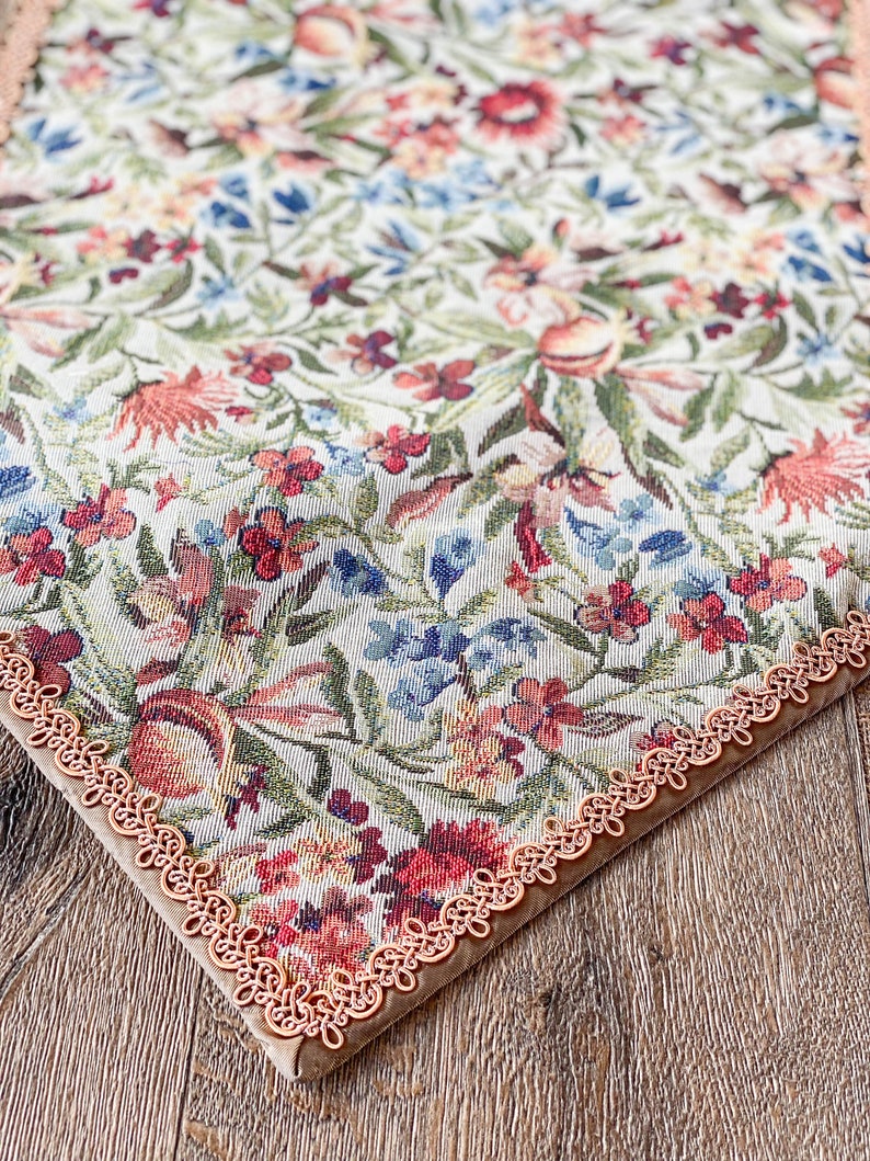 Pode incluir: Um corredor de mesa floral com uma borda de renda bege. O corredor de mesa apresenta um fundo branco com um padr&atilde;o repetitivo de flores vermelhas, rosas, azuis e verdes.