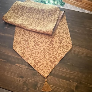 Può includere: Runner da tavolo color oro con un motivo floreale ornato e ripetuto. Il runner è realizzato in un tessuto strutturato e ha un nappa abbinata all'estremità. Il runner è piegato su una superficie di legno scuro.