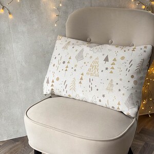 May include: A white chair with a cream-colored cushion and a throw pillow with a gold and brown Christmas tree pattern. The chair is in front of a wall with string lights.
