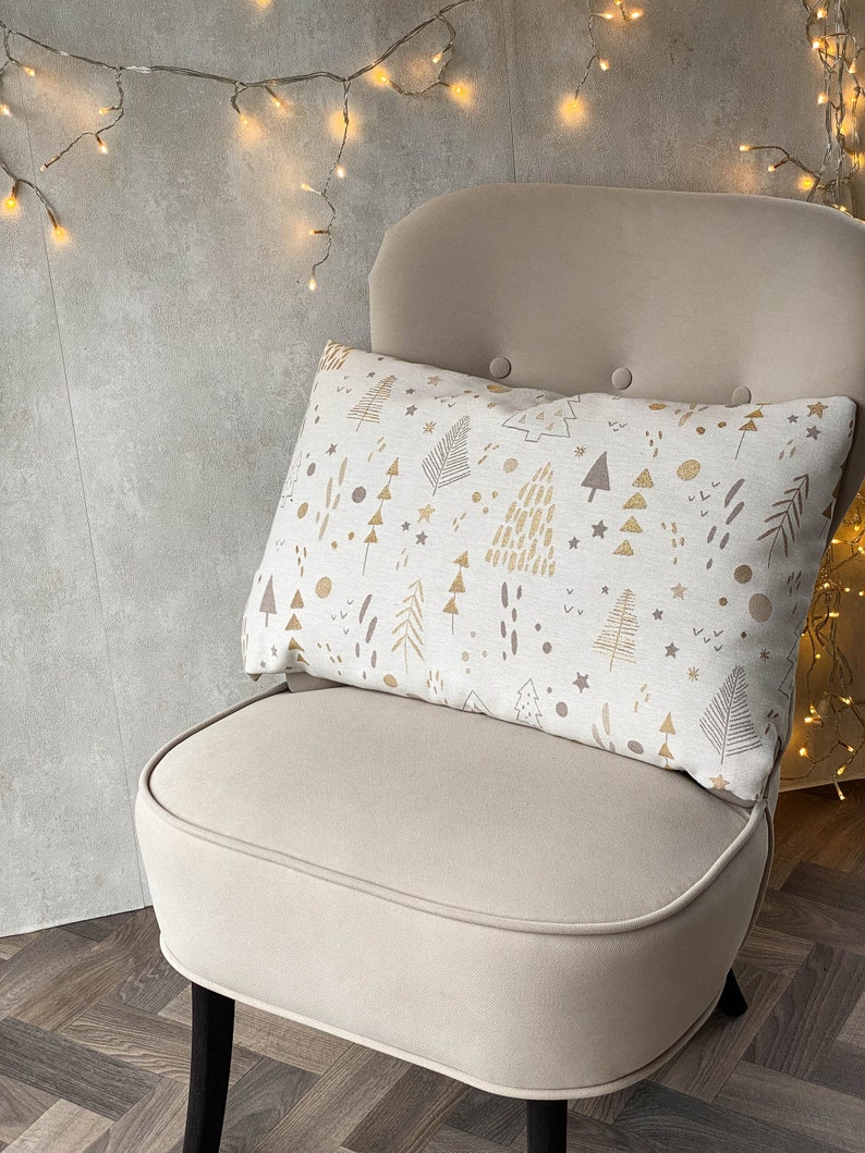 May include: A white chair with a cream-colored cushion and a throw pillow with a gold and brown Christmas tree pattern. The chair is in front of a wall with string lights.