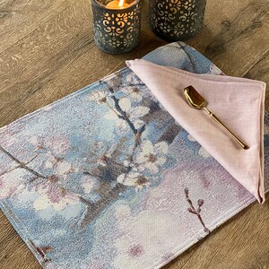 May include: A blue and white floral patterned placemat with a pink linen napkin and a gold spoon on top. The placemat is on a wooden table with two candle holders.