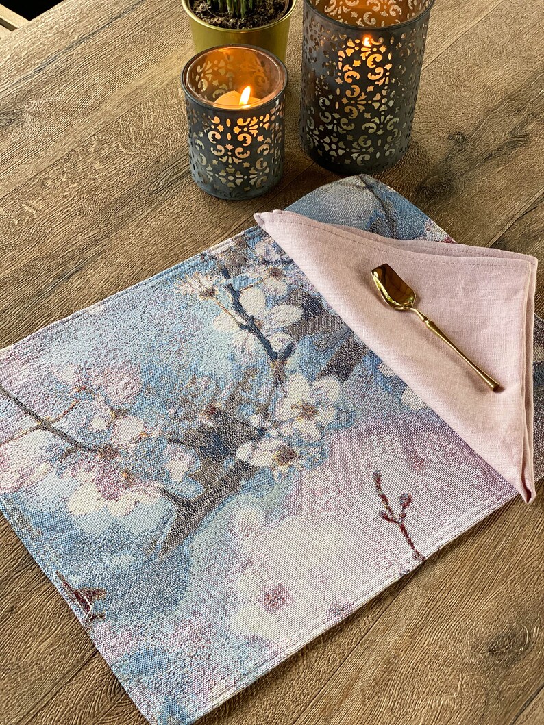 May include: A blue and white floral patterned placemat with a pink linen napkin and a gold spoon on top. The placemat is on a wooden table with two candle holders.