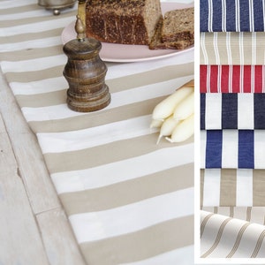 May include: A beige and white striped table runner with a wooden pepper mill, a plate of sliced bread, and a bunch of white candles. The table runner is shown on a whitewashed wooden surface.