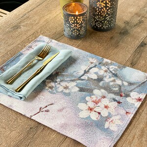 May include: A blue and white floral placemat with a light blue napkin and gold silverware on top. Two decorative candle holders are in the background.