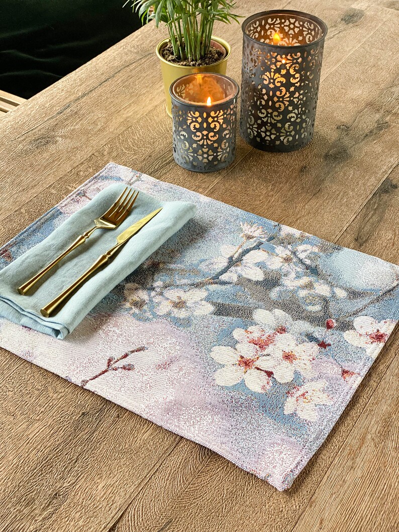 May include: A blue and white floral placemat with a light blue napkin and gold silverware on top. Two decorative candle holders are in the background.