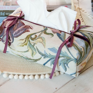 May include: A floral patterned tissue box cover with a burgundy ribbon tied around it. The tissue box cover is sitting on a round, cream-coloured woven mat with white pom-poms around the edge.