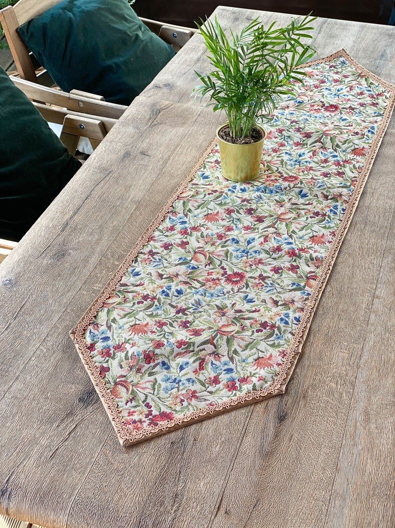 Pode incluir: Um corredor de mesa floral com uma borda festonada e um fundo bege. O corredor tem um padr&atilde;o floral em tons de vermelho, azul e verde. Uma pequena planta em vaso est&aacute; em cima do corredor de mesa.