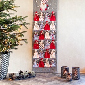 Puede incluir: Un calendario de adviento navide&ntilde;o de fieltro gris con 24 bolsillos de tela roja y blanca. Un gnomo con barba blanca y gorro rojo est&aacute; en la parte superior del calendario. El calendario est&aacute; colgado en una pared blanca.