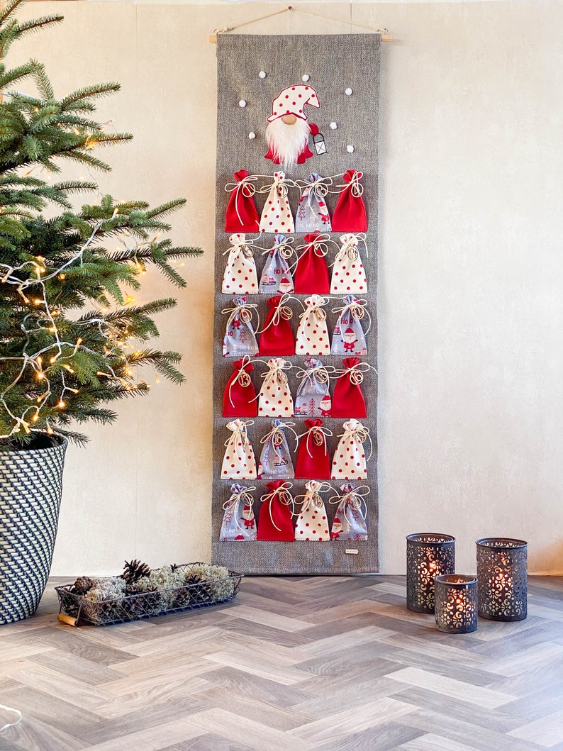 Puede incluir: Un calendario de adviento navide&ntilde;o de fieltro gris con 24 bolsillos de tela roja y blanca. Un gnomo con barba blanca y gorro rojo est&aacute; en la parte superior del calendario. El calendario est&aacute; colgado en una pared blanca.