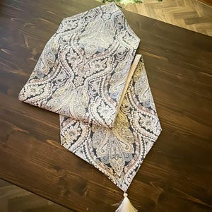 Può includere: Runner da tavolo con motivo paisley nei toni del nero, crema e azzurro. Il runner ha un nappa decorativa all'estremità. Il runner è posto su un tavolo di legno marrone scuro.