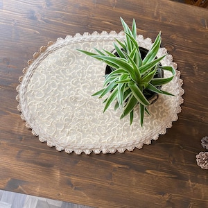 May include: A beige oval table runner with a lace trim and a delicate floral pattern. A potted green plant with white stripes sits on top of the runner.
