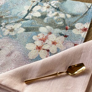 May include: A pink linen napkin with a gold spoon on top. The napkin is folded in half and the spoon is lying on top of the napkin. The spoon is a traditional style with a rounded bowl and a long handle. The napkin is a light pink color and has a soft, textured surface. The spoon is a shiny gold color and has a smooth surface.