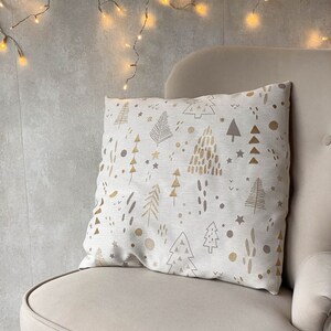 May include: A beige throw pillow with a gold and brown pattern of stylized Christmas trees, stars, and dots. The pillow is sitting on a beige armchair with a string of white Christmas lights hanging above it.