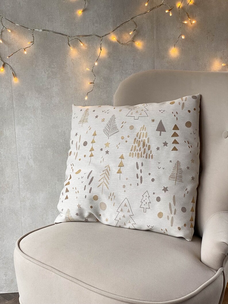 May include: A beige throw pillow with a gold and brown pattern of stylized Christmas trees, stars, and dots. The pillow is sitting on a beige armchair with a string of white Christmas lights hanging above it.