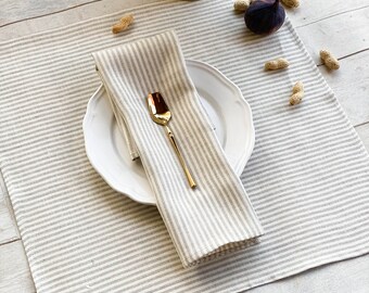 May include: A white and gray striped linen placemat with a white plate and a gold spoon on top. The placemat is on a white wooden table with peanuts and a fig scattered around.