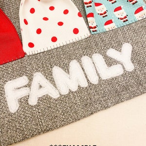 Puede incluir: Un calendario de adviento de arpillera gris con bolsillos de fieltro blanco y la palabra "FAMILY" en letras de fieltro blanco. Los bolsillos est&aacute;n decorados con lunares rojos y blancos, patrones de Pap&aacute; Noel y un adorno de &aacute;rbol de Navidad de madera.