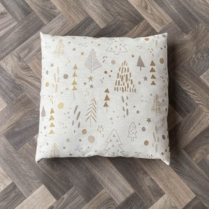 May include: A white throw pillow with a gold and gray Christmas tree pattern. The pillow is on a wood floor.