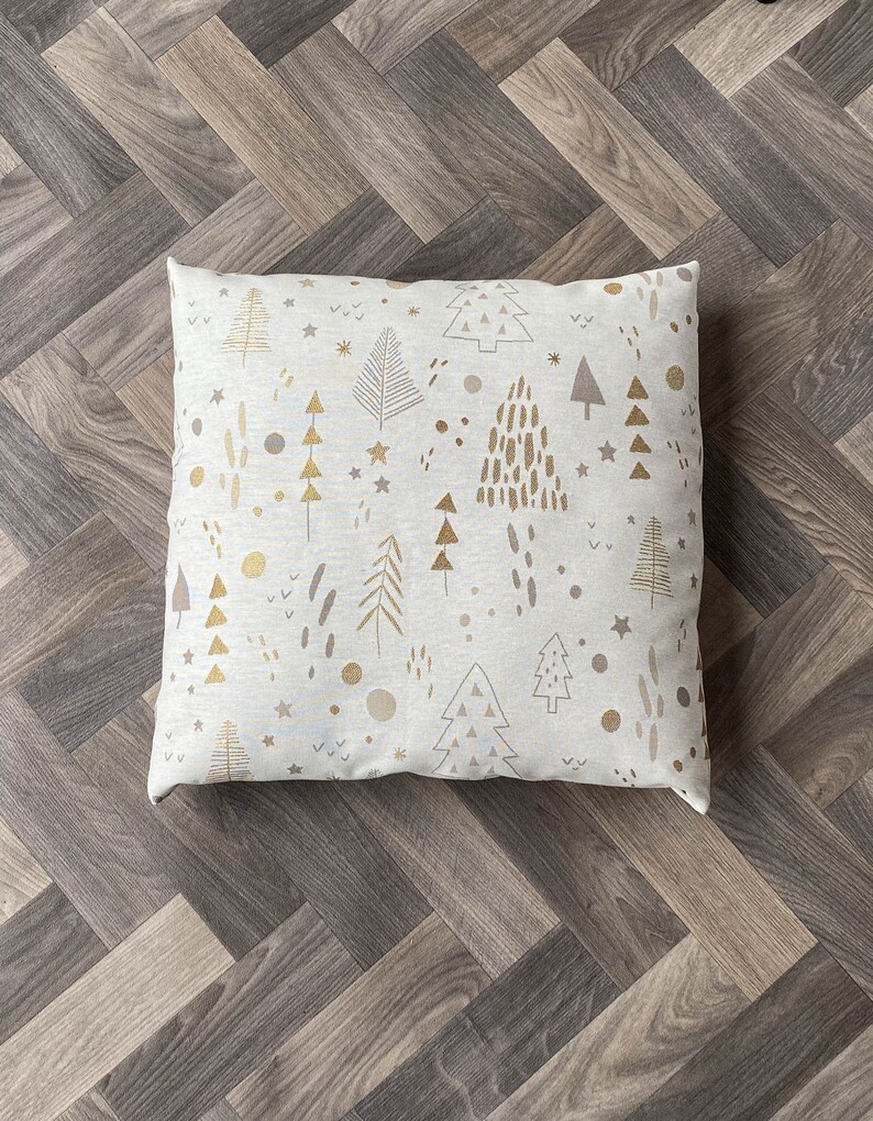 May include: A white throw pillow with a gold and gray Christmas tree pattern. The pillow is on a wood floor.