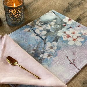 May include: A blue and pink floral placemat with a gold spoon on a pink linen napkin. The placemat has a woven texture and features a delicate floral design.