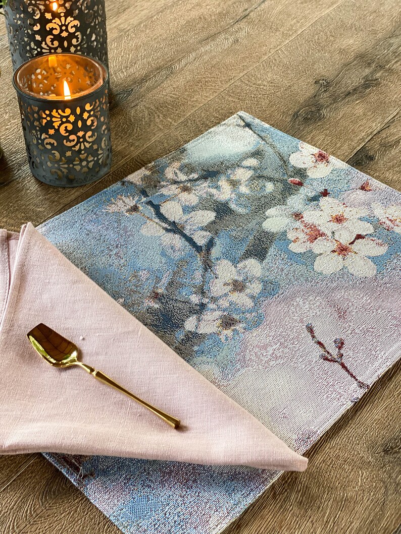 May include: A blue and pink floral placemat with a gold spoon on a pink linen napkin. The placemat has a woven texture and features a delicate floral design.