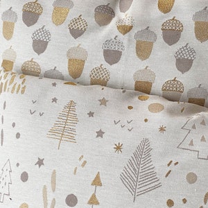 May include: Two decorative throw pillows with a white background and gold and brown patterns. The top pillow has a repeating acorn pattern. The bottom pillow has a repeating pattern of stylized trees, stars, and dots.