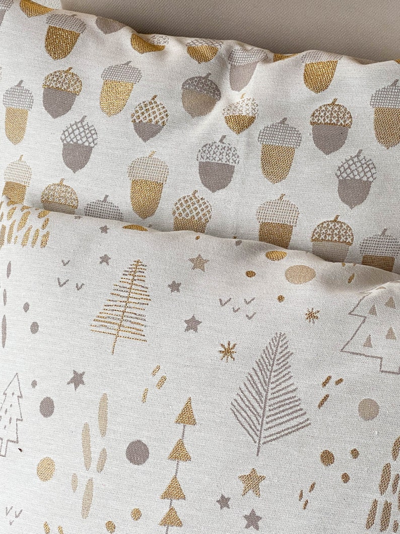 May include: Two decorative throw pillows with a white background and gold and brown patterns. The top pillow has a repeating acorn pattern. The bottom pillow has a repeating pattern of stylized trees, stars, and dots.