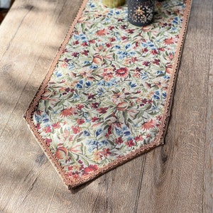 Pode incluir: Um corredor de mesa floral com uma borda festonada e um fundo bege. O corredor apresenta uma variedade de flores em tons de vermelho, rosa, azul e verde. O corredor est&aacute; colocado sobre uma mesa de madeira.