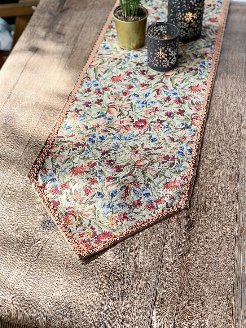 Pode incluir: Um corredor de mesa floral com uma borda festonada e um fundo bege. O corredor apresenta uma variedade de flores em tons de vermelho, rosa, azul e verde. O corredor est&aacute; colocado sobre uma mesa de madeira.